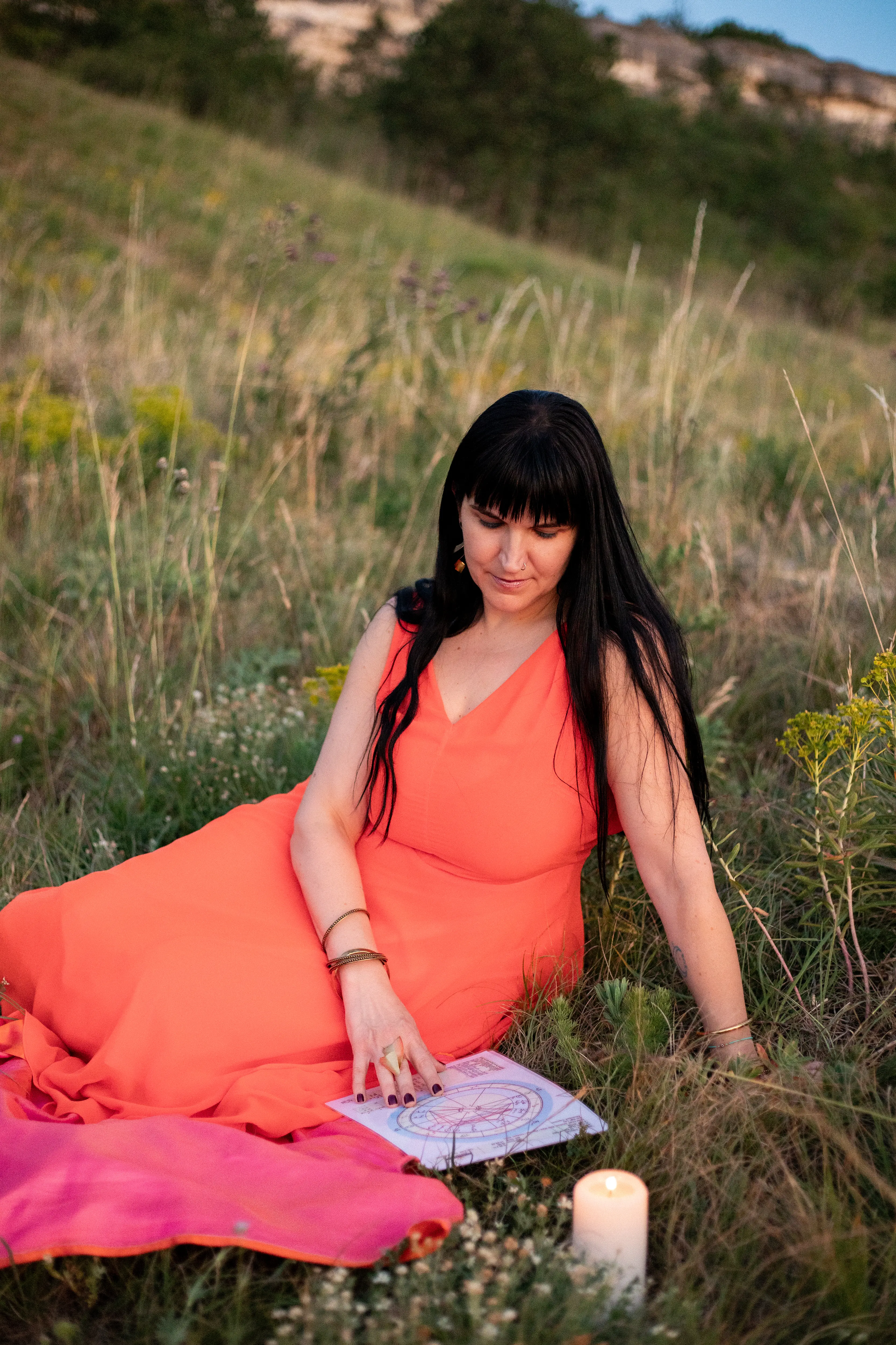 Adrienne sitting in a sunlit field with a printed natal chart and a lit candle