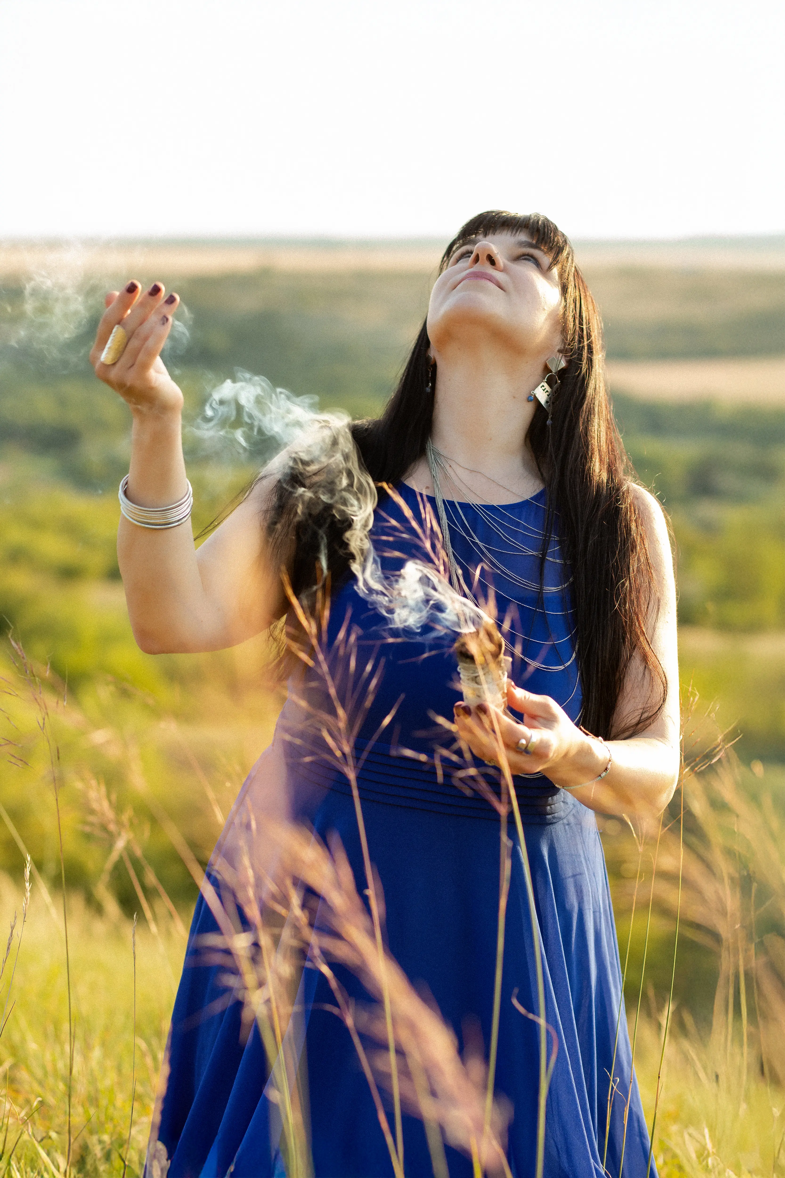 Adrienne looking up to the sky with sage smoke rising around her in a sunlit field